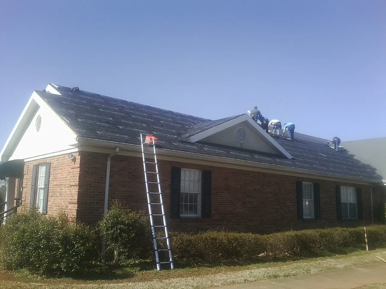 Crew tearing shingles off a brick ranch roof with a ladder set — Emergency Roof Repair work by our crew in Locust Hill