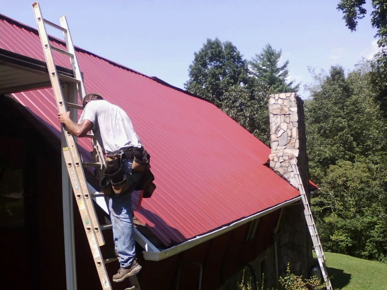 Roofer on a ladder beside a red metal roof and stone chimney — Soffit & Fascia Repair by Roofing Brothers in Locust Hill